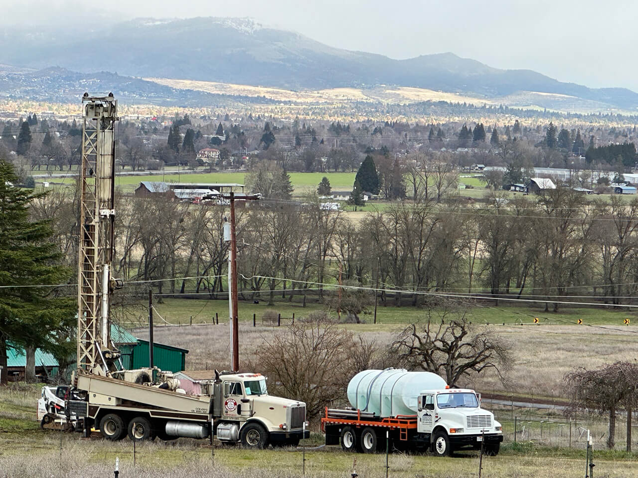 A photo showing the Siskiyou mountains in the background and 2 water well drilling trucks and rig in the foreground